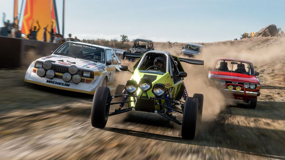 A group of off-road cars including a green buggy, red half-truck and white rally car drive along a dirt trail with spectators cheering on the action in the left side of the image.