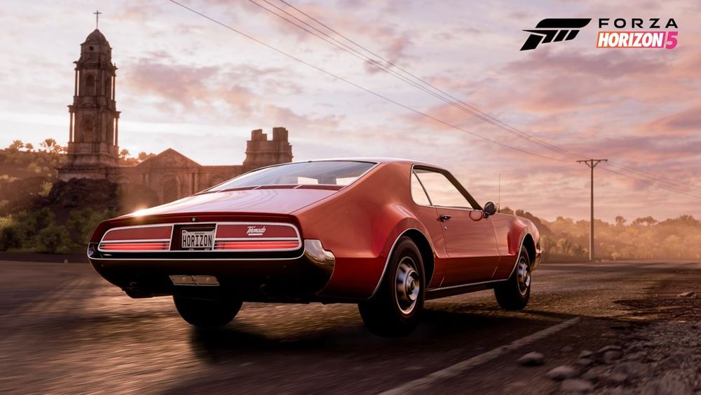 An orange Oldsmobile Toronado by a Mexican architectural site is parked on the road as a pink sky sets in after sunset.