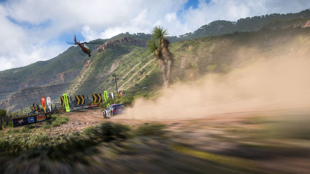 Colin McRae's Ford Focus RS kicks up dust as it blasts down a dirt trail in Sierra Nueva, surrounded by rocky terrain covered in greenery.