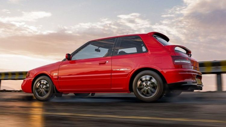 A red Mazda 323 GTR drives along a wet surface road during sunrise.