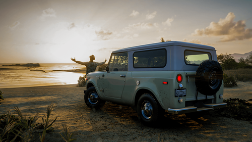 A player stands in front of their car facing the sunrise on a beach, with their arms outstretched as if to welcome it.