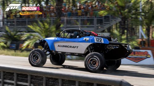 An Alumicraft buggy with a blue and white livery drives past a grandstand with spectators in the Palm Forest.