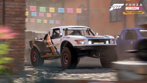 The Alumicraft Trick Truck drives through a town side by side with a blue Fastball Racing truck. Orange and red brick buildings can be seen behind them.