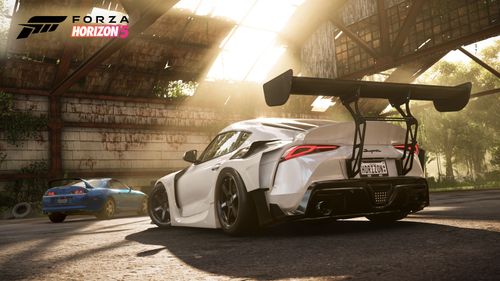 Two eras of the Toyota Supra, the modern car white and fitted with a body kit, the legendary classic painted in blue, parked in an abandoned airfield hangar.