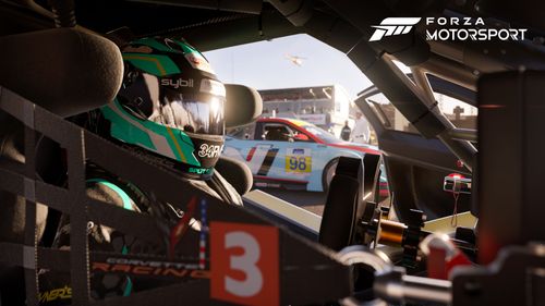 A driver in a cyan coloured race suit and helmet sitting in the 2020 Chevrolet #3 Corvette Racing C8.R with the door open. A blue and red  2020 Hyundai #98 Bryan Herta Autosport Veloster N can also be seen in the background.